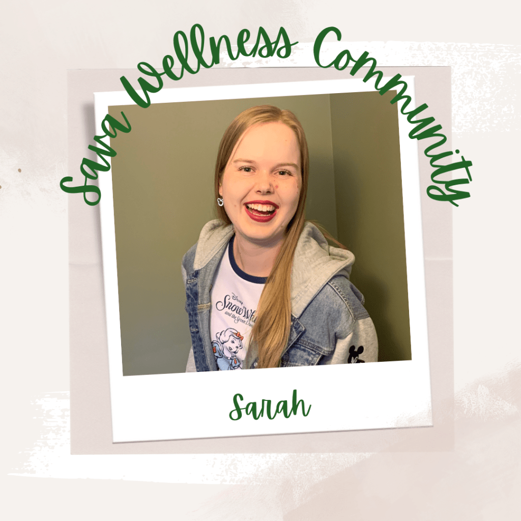 The words āSava Wellness Communityā are written in green cursive script over top of a polaroid picture. The polaroid picture is sitting on top of multiple layers of pink, cream, and white watercolour background. The polaroid picture has the name āSarahā written in green under it. The picture is of Sarah smiling at the camera. Sarah has her blonde hair down, is wearing red lipstick, and has a jean jacket over a t-shirt on.