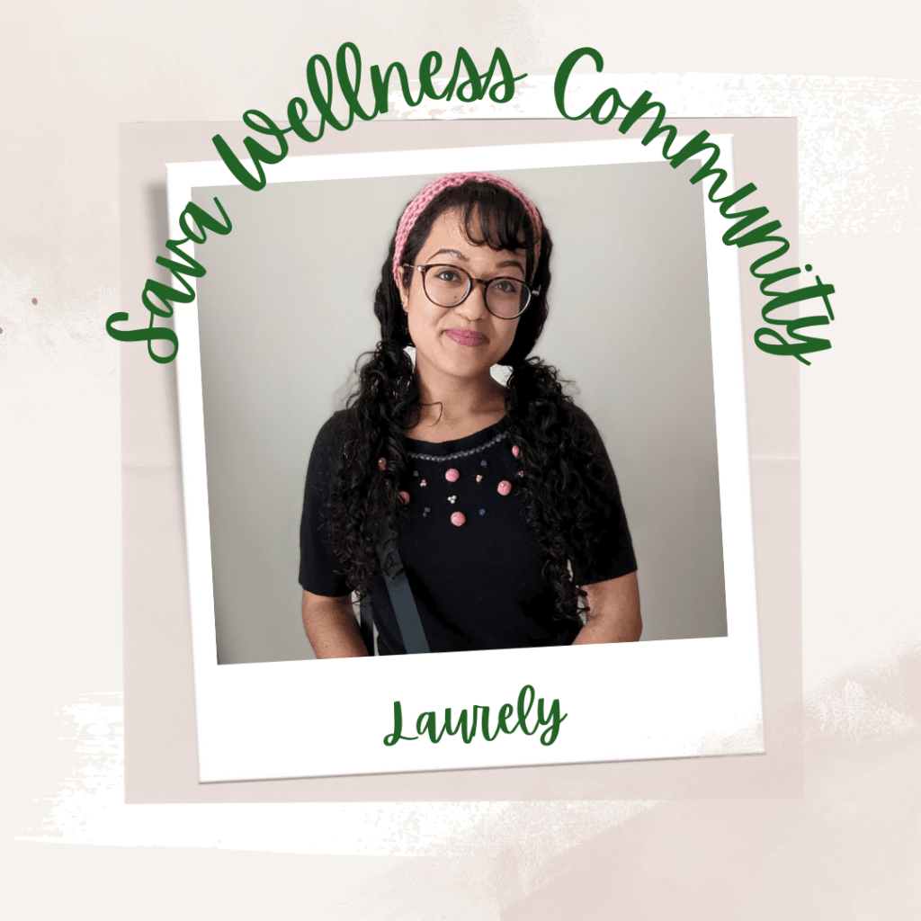 The words “Sava Wellness Community” are written in green cursive script over top of a polaroid picture. The polaroid picture is sitting on top of multiple layers of pink, cream, and white watercolour background. The polaroid picture has the name “Laurely” written in green under it. The picture is of Laurely smiling at the camera with her hair in pigtails and a pink crocheted headband. Laurely wears glasses and is wearing a black t-shirt with crochet elements and has a bag over her right shoulder.