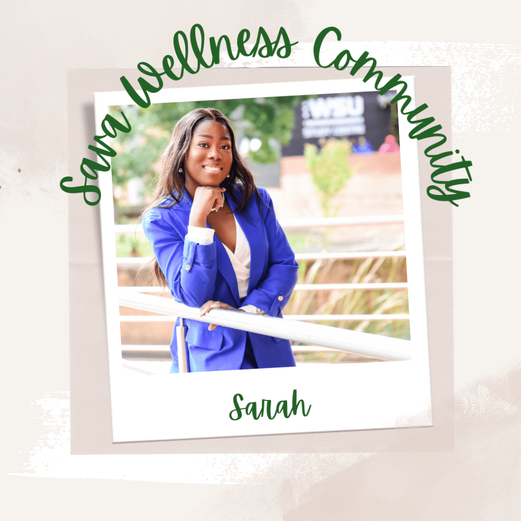 The words “Sava Wellness Community” are written in green cursive script over top of a polaroid picture. The polaroid picture is sitting on top of multiple layers of pink, cream, and white watercolour background. The polaroid picture has the name “Sarah” written in green under it. The picture is of Sarah in a blue blazer with a white shirt underneath. Sarah is leaning on a white railing, with her chin resting on her right arm. Sarah is smiling at the camera.