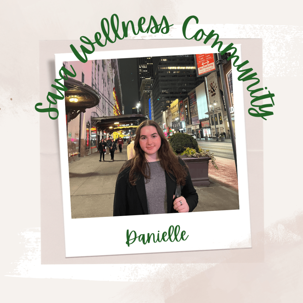 The words “Sava Wellness Community” are written in green cursive script over top of a polaroid picture. The polaroid picture is sitting on top of multiple layers of pink, cream, and white watercolour background. The polaroid picture has the name “Danielle” written in green under it. Danielle is looking at the camera from a city street at nighttime. She is wearing a grey sweater, a black blazer, and has her left hand holding her purse. She has brown hair and is wearing a metal ring on her finger.